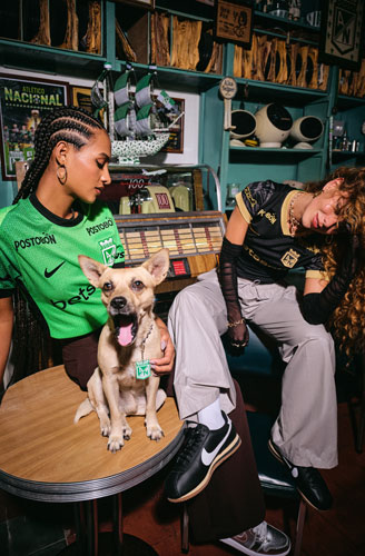 Mujer con la camiseta verde del tercer kit y otra mujer con la camiseta negra de visitante de Atlético Nacional 2025, junto a un perro, en un bar deportivo.
